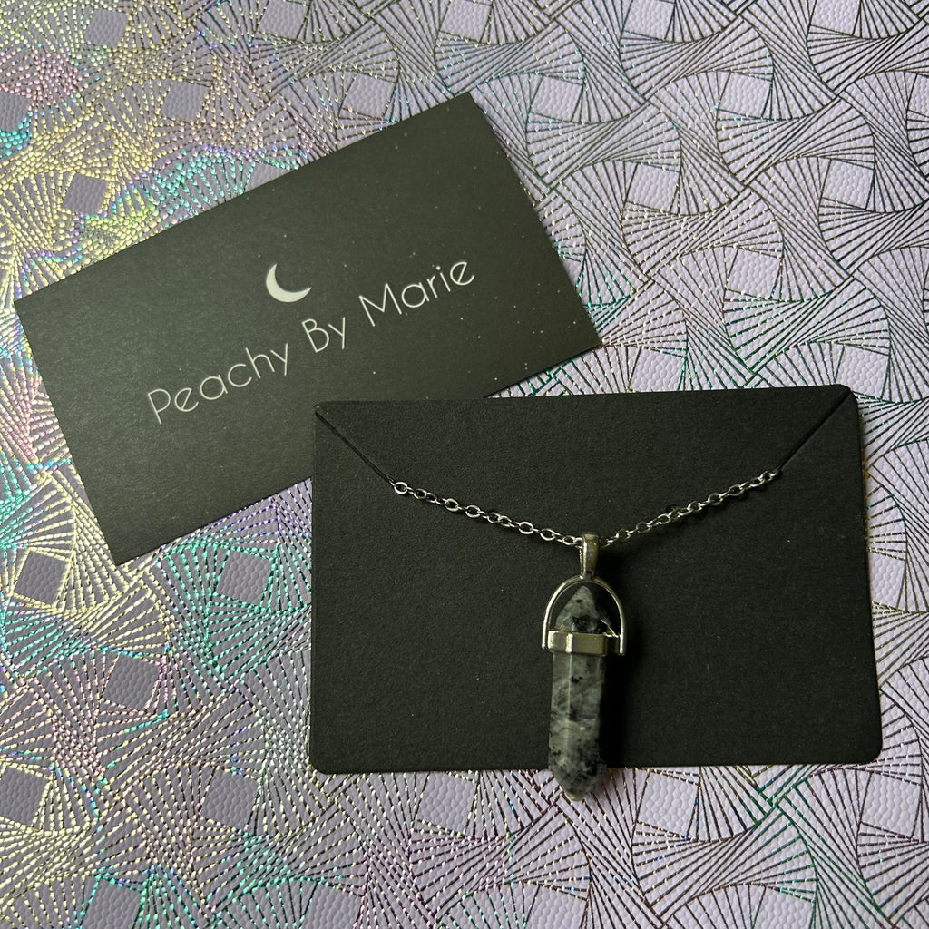 Black crystal necklace, the crystal is long and has six sides, pointy on the top and bottom.  There is a metal clasp holding the crystal, and a fine stainless steel chain with a small clasp. The picture also depicts the brand card from Peachy By Marie.